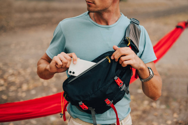 Ένας νεαρός άνδρας με ένα T-shirt και μια τσάντα μέσης στον ώμο του βάζει ένα smartphone στην τσέπη του, κοντά - Φωτογραφία, εικόνα
