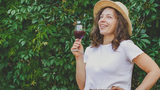 Νεαρή όμορφη γυναίκα που κρατά ένα ποτήρι κόκκινο κρασί, χαμογελώντας χαλαρώνοντας κοιτάζοντας σε απόσταση, φορώντας καλοκαιρινό ψάθινο καπέλο και λευκό t-shirt, στέκεται στο πράσινο πάρκο ή κήπο έξω από την πόρτα. - Φωτογραφία, εικόνα