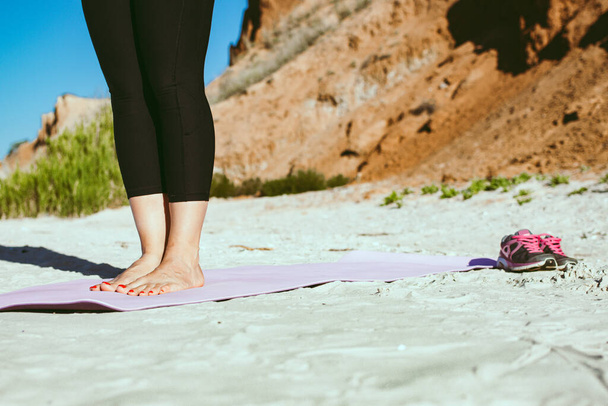 ビーチでヨガ。屋外でストレッチ足のワークアウトを行う前に、スポーツマットの上に立つ裸足の女性の足、黒のスポーツウェアを着て女性は、スニーカーは砂の上に立つ。健康的な趣味の幸福の概念 - 写真・画像