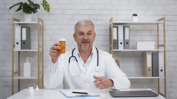 Médico maduro mostrando tubo con píldoras médicas sentado en la oficina - Imágenes, Vídeo