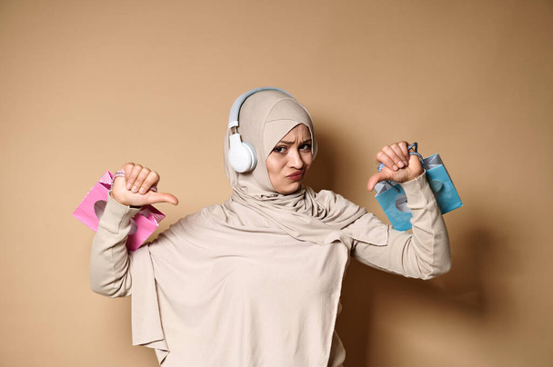 Zdenerwowana kobieta w hidżabie i słuchawki trzymające w dłoniach różowe i niebieskie torby z papierem upominkowym i pokazujące opuszczone kciuki, wyrażające niezadowolenie - Zdjęcie, obraz