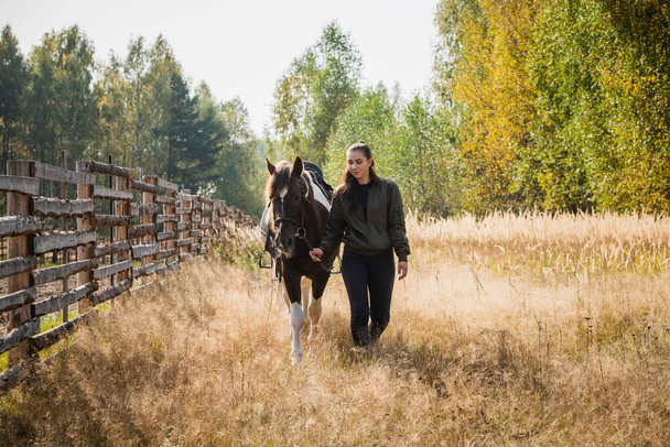 Caminar a una chica con un caballo a lo largo de la valla en el rancho en el otoño - Foto, imagen