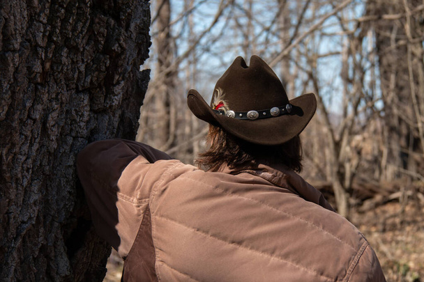 Een vrouw met kort bruin haar draagt een bruine ridge top cattleman cowboy hoed met een veer en decoratieve riem bevestigd. Leunend tegen een boom op een boerderij in de buurt van Londen, Ontario, Canada, voorjaar 2021.  - Foto, afbeelding