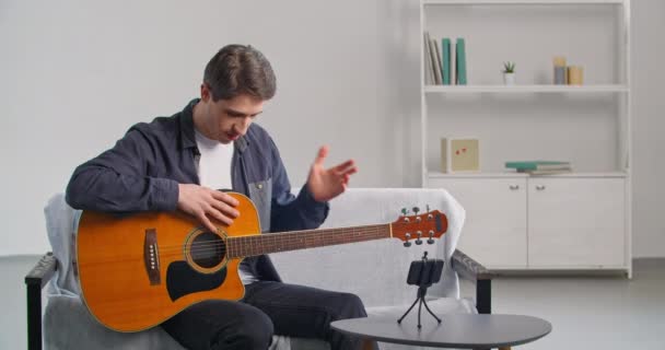 Νεαρός δάσκαλος με casual ρούχα ταλαντούχος άνδρας καλλιτέχνης κάθεται στον καναπέ στο σπίτι κάνοντας online μάθημα webinar διδασκαλία βίντεο σχετικά με το παιχνίδι κιθάρα δείχνει δομή του μουσικού οργάνου εξηγεί - Πλάνα, βίντεο