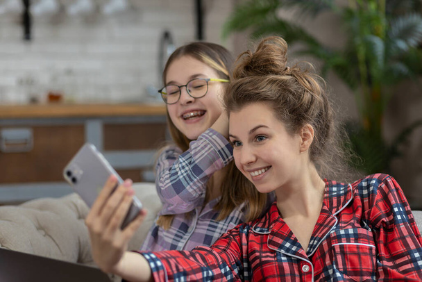 Duas adolescentes de pijama fazem uma chamada de selfie ou vídeo enquanto estão sentadas no sofá em casa - Foto, Imagem