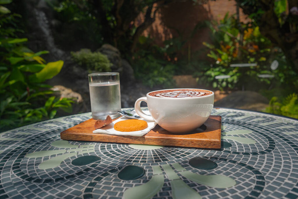 Een set warme koffiedranken geplaatst op een keramische tafel. De achtergrond van een waterval, weelderig groen en stoom dreef uit om het er fris uit te laten zien - Foto, afbeelding