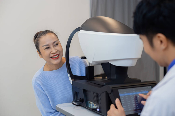 診療所の目の専門家による近代的な視力測定機器を検討するアジアのシニア女性。眼科医の診断コンセプト. - 写真・画像