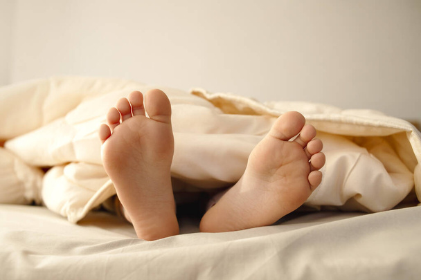 Os pés de bebé das raparigas espreitam debaixo do cobertor. Pernas saindo da cama, deitadas no hotel em casa. - Foto, Imagem