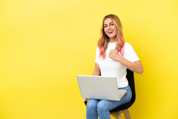 Dizüstü bilgisayarlı bir sandalyede oturan genç bir kadın izole edilmiş sarı arka planda baş parmak hareketi yapıyor. - Fotoğraf, Görsel