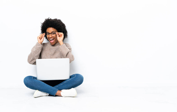 Joven mujer afroamericana con una laptop sentada en el suelo con gafas y sorprendida - Foto, imagen