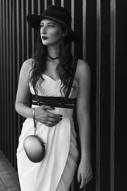 Young beautiful sexy woman wearing trendy outfit, white dress, black hat and leather swordbelt. Longhaired brunette posing in the city street. Outdoor fashion photography. - Photo, Image
