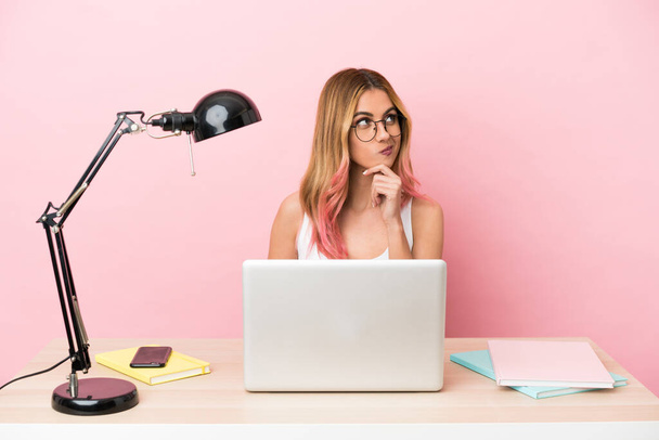 Νεαρή φοιτήτρια σε ένα χώρο εργασίας με ένα φορητό υπολογιστή πάνω από ροζ φόντο σκέφτεται μια ιδέα, ενώ ψάχνει επάνω - Φωτογραφία, εικόνα