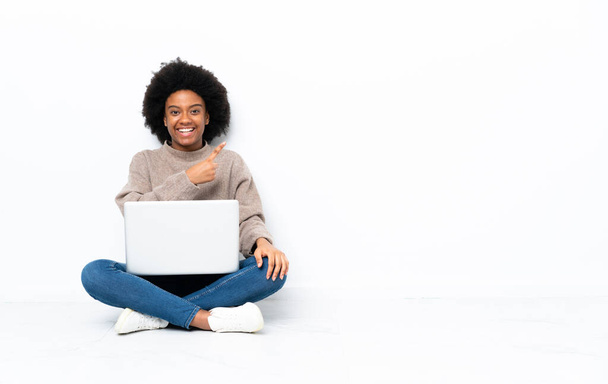 Молода афроамериканська жінка з ноутбуком сидить на підлозі, вказуючи на бік, щоб представити продукт. - Фото, зображення