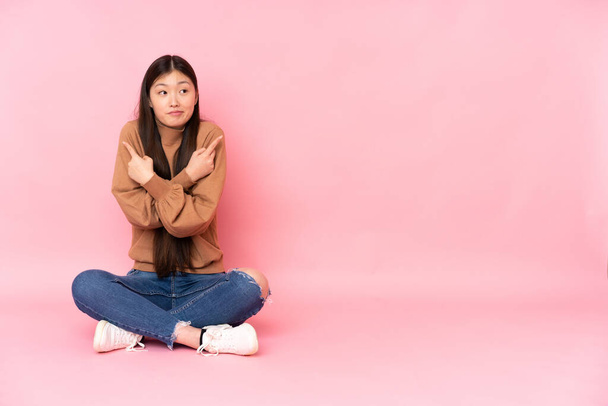 Młoda Azjatka siedząca na podłodze odizolowana na różowym tle wskazując na strony mające wątpliwości - Zdjęcie, obraz