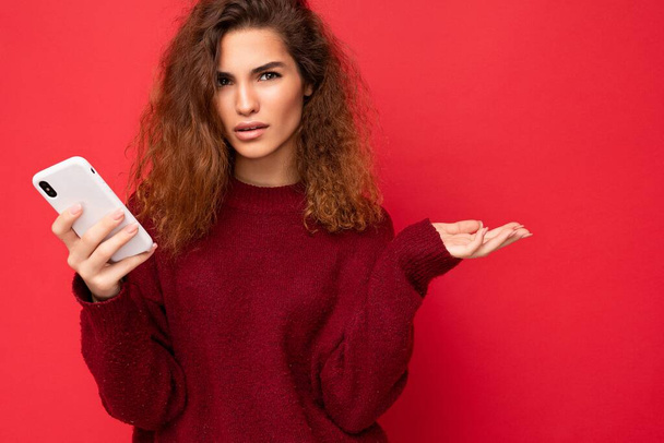 Close up Photo of pretty serious adult woman with curly hair wearing dark red sweater isolated on red background holding mobile phone looking at camera and asking - Photo, Image