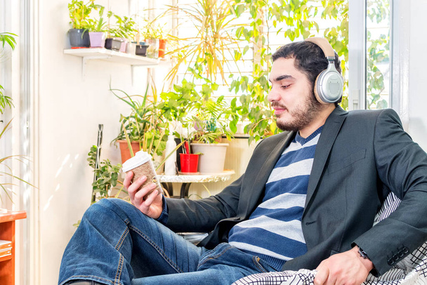 A young Caucasian bearded man listening to music with wireless headphones, holding a cup of coffee - Foto, Bild