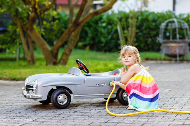 Mała dziewczynka maluch bawi się dużym zabytkowym samochodem zabawki i zabawy na świeżym powietrzu w lecie. Ładne dziecko zatankować samochód wodą. Dziewczyna za pomocą węża ogrodowego i wypełnić benzyną, gra roli stacji benzynowej. - Zdjęcie, obraz