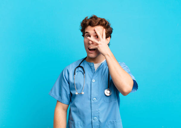 young nurse man looking shocked, scared or terrified, covering face with hand and peeking between fingers - Photo, image