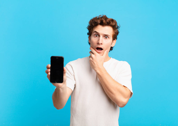 young boy with mouth and eyes wide open and hand on chin, feeling unpleasantly shocked, saying what or wow. telephone screen concept - Photo, Image