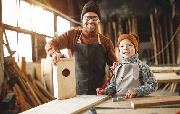 Hombre barbudo adulto alegre e hijo pequeño en delantales que ensamblan la casa de madera del pájaro juntos mientras que trabajan en taller de carpintería - Foto, Imagen