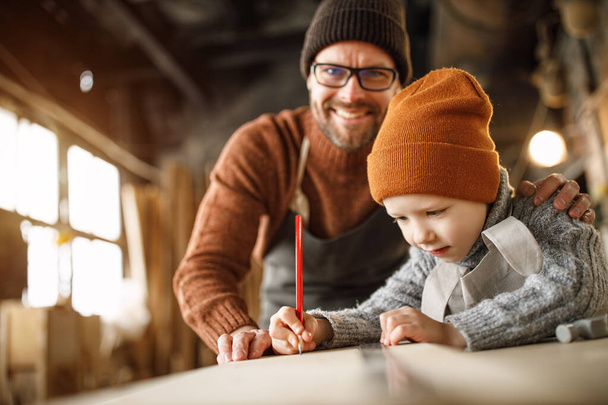 Excited little boy drawing sketch on wooden plank with help of father while spending day with dad in carpentry workshop - Photo, Image