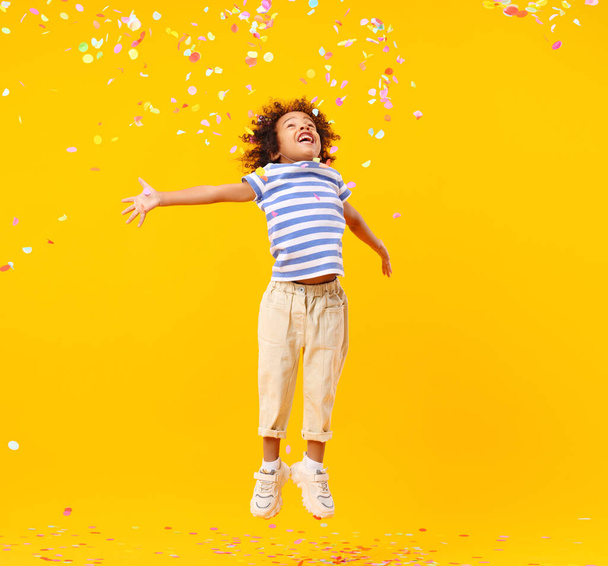 Alegre niño negro con el pelo afro riendo y saltando al tratar de atrapar confeti colorido durante la celebración del cumpleaños contra el fondo amarillo - Foto, imagen