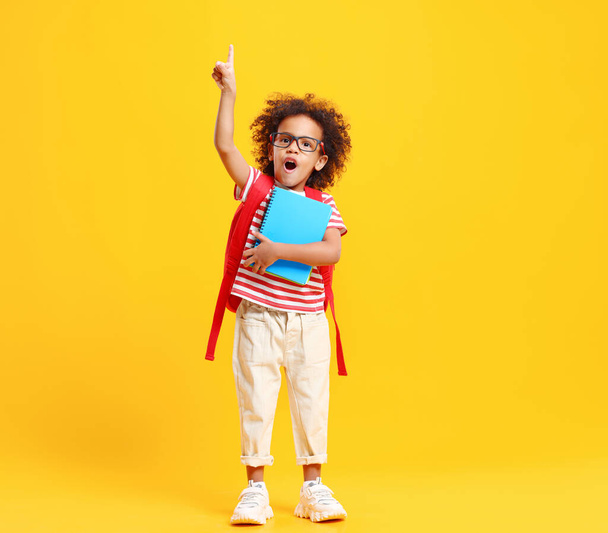 Cheerful smart African American little schoolboy with curly hair in casual clothes and eyeglasses  looking at camera while having idea standing against yellow background with notebooks - Fotoğraf, Görsel