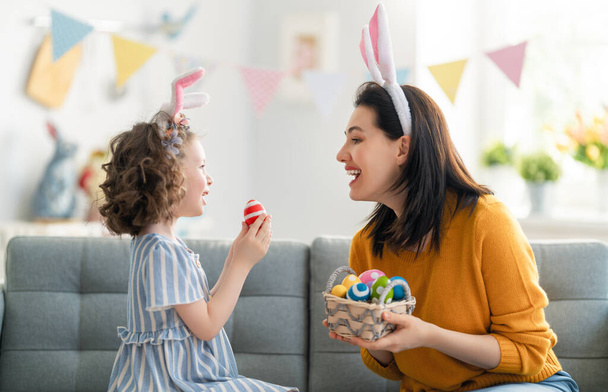 Happy holiday! Mother and her daughter with painting eggs. Family celebrating Easter. Cute little child girl is wearing bunny ears.  - Foto, afbeelding