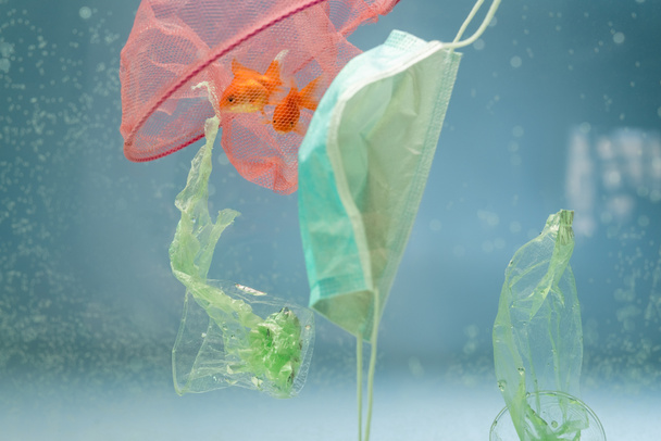red con peces de colores, máscara médica, bolsas de plástico y tazas en agua, concepto de ecología - Foto, imagen