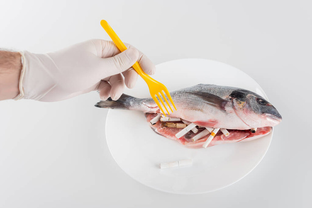 partial view of male hand in latex glove, with plastic fork, near fish stuffed with cigarette ends on white, ecology concept - Photo, Image