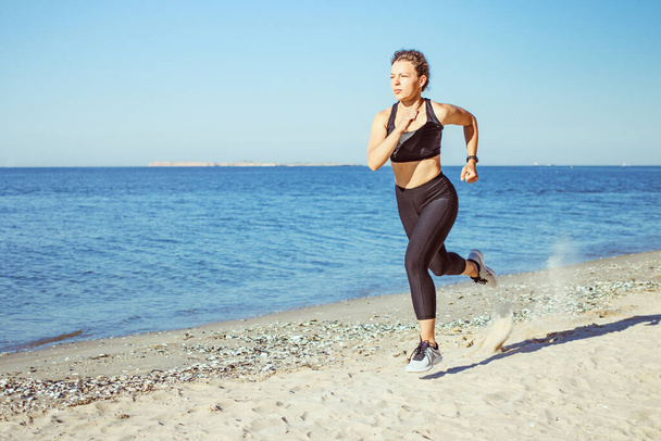 Concentrated young woman jogger running on beach outdoor workout in morning, wearing black sportswear on blue sea and sky background. Weight loss cardio goal achievement challenge. Copy space. - Photo, Image
