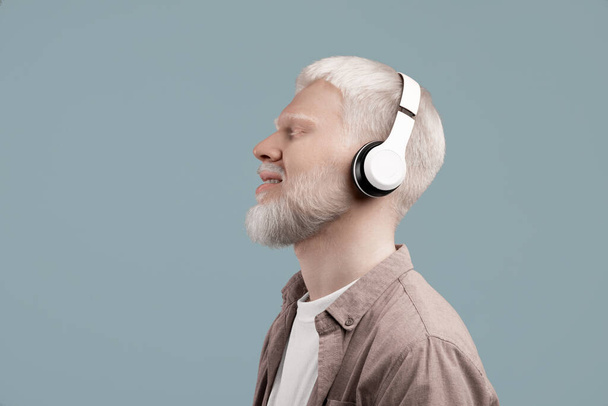 Spokojny albinos korzystający z muzyki poprzez bezprzewodowe słuchawki z zamkniętymi oczami i uśmiechem, słuchając ulubionych melodii lub medytacji audio - Zdjęcie, obraz