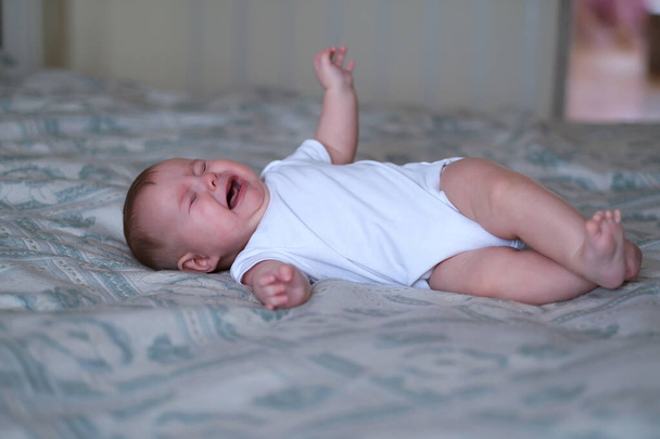 Pullukka söpö sinisilmäinen vauva valkoisessa kehonpuvussa sängyllä itkee, nyyhkii, huutaa - Valokuva, kuva