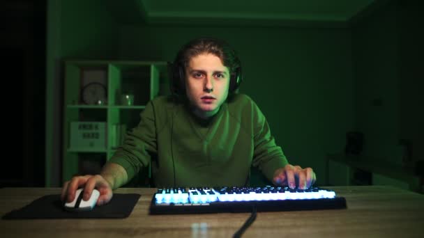 Gericht gamer met een gespannen gezicht speelt videospelletjes thuis op een computer in een kamer met groene lichten en kijkt boos naar de camera. Emotionele man in een headset speelt online games en streaming. - Video