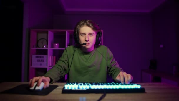 Positieve gamer in een headset thuis zitten 's nachts op de computer en typen op het toetsenbord met een glimlach op zijn gezicht te kijken naar de camera. - Video