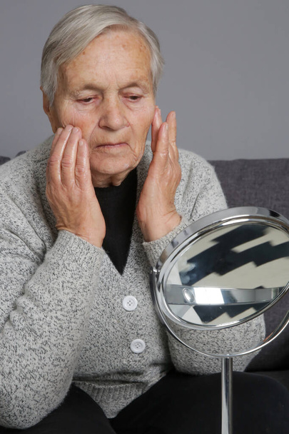 Oudere vrouw met gerimpelde huid met behulp van huidverzorgingsproducten in de voorkant van de spiegel thuis. Huiselijk leven, spa, zelfverzorging en verwennerij. - Foto, afbeelding