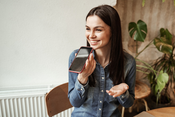 Lächelnde junge Frau nimmt Sprachnachricht auf, während sie ihr Smartphone in der Hand hält  - Foto, Bild