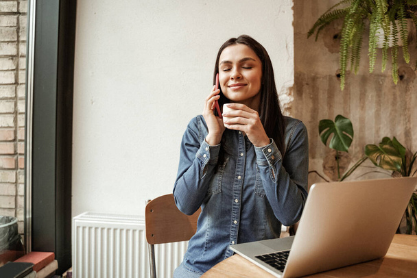 femme heureuse tenant tasse de café tout en parlant sur smartphone près d'un ordinateur portable - Photo, image