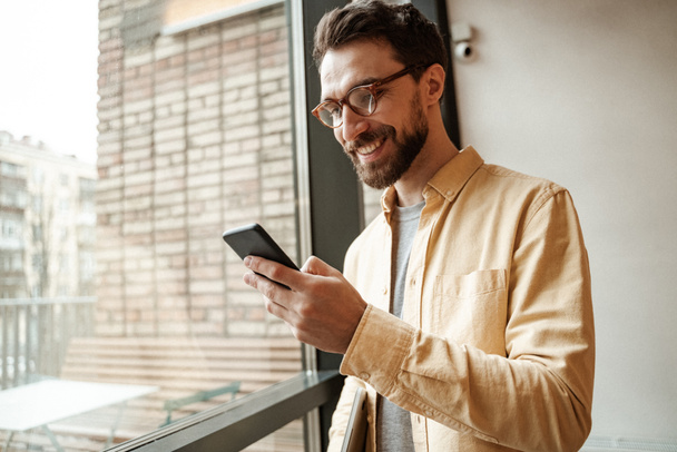 smiling and bearded man using smartphone while standing near window  - Photo, Image