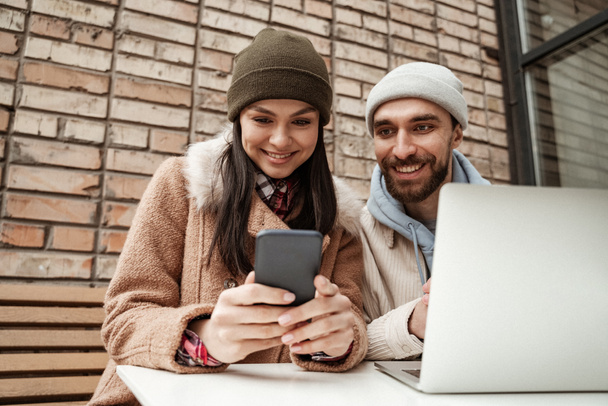 happy freelancers in beanie hats looking at smartphone near laptop on table - Photo, Image