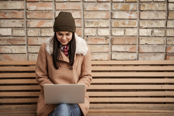 szczęśliwy brunetka freelancer w czapka kapelusz za pomocą laptopa podczas siedzi na ławce - Zdjęcie, obraz