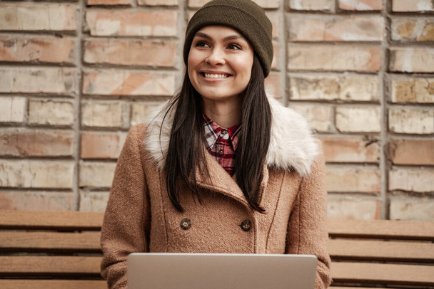 usmívající se brunetka na volné noze v čepici pomocí notebooku, zatímco sedí na lavičce - Fotografie, Obrázek