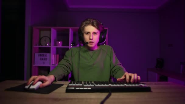 Спокійний концентрований молодий чоловік в гарнітурі грає в відеоігри вдома вночі в кімнаті з фіолетовим світлом
. - Кадри, відео