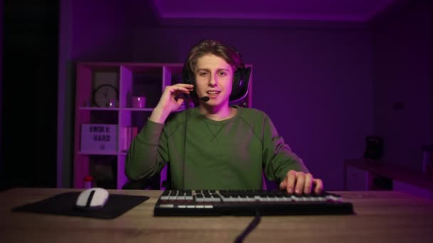 Joven guapo comienza a transmitir juegos de ordenador en línea en casa por la noche, se comunica en un auricular y sonríe. - Imágenes, Vídeo