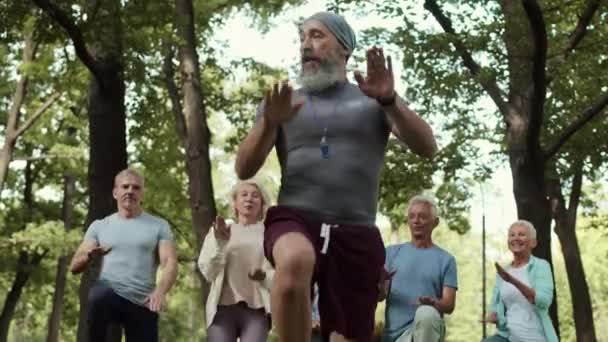 Μέτριο πλάνο γενειοφόρος γέρος εκπαιδευτής φυσάει σφυρίχτρα και στη συνέχεια να αρχίσει να τρέχει στη θέση του με αρκετούς ηλικιωμένους αθλητικούς ανθρώπους πίσω του στο πάρκο - Πλάνα, βίντεο