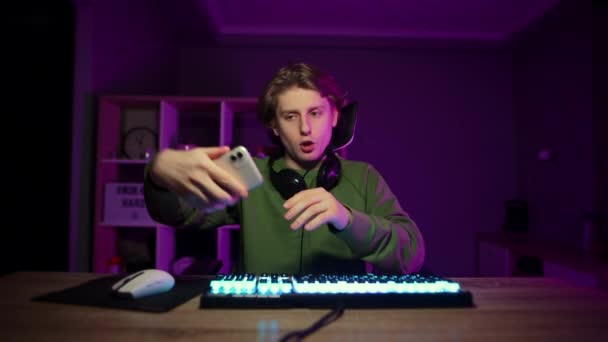 Jonge gamer in een headset zit 's nachts achter een computer in een kamer en selfie op smartphone camera, toont een gebaar van vrede. - Video