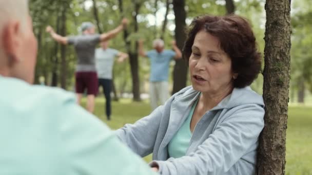 Vue arrière de belle femme sportive âgée assise sur la pelouse dans le parc et bavardant avec une femme méconnaissable avec une coupe de cheveux courte assise devant elle - Séquence, vidéo