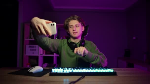 Jugador en un auricular se sienta en una computadora por la noche en una habitación y hace una videollamada en un teléfono inteligente, muestra un gesto de saludo con la palma de su mano y sonríe. - Imágenes, Vídeo