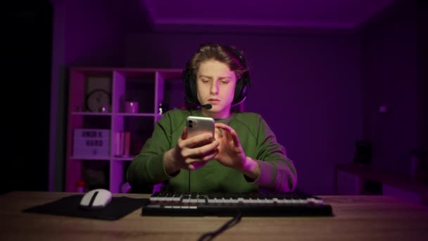 Enfocado joven se sienta por la noche en una habitación con una luz púrpura detrás de un ordenador y utiliza un teléfono inteligente con una cara pensativa. Gamer utiliza un teléfono inteligente durante el descanso después del juego. - Imágenes, Vídeo