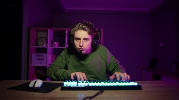 Un jugador pensativo con auriculares en la cabeza usa una computadora con una cara seria. Chico de noche en el dormitorio juega juegos de ordenador. - Metraje, vídeo